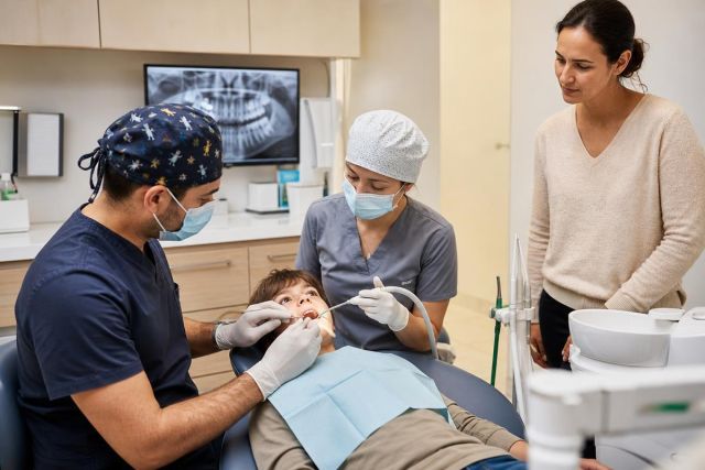 Pediatric dental exam in clinic: dentist and hygienist treating child while parent observes.