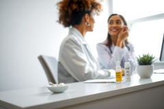 Doctor consults with a patient in a bright, modern clinic. A white desk holds medication and a plant. The scene emphasizes healthcare and patient care.
