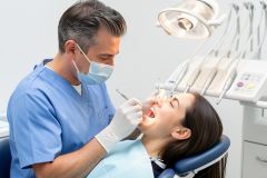 Dentist in mask examines a patient's open mouth at a dental chair, assessing gums and periodontal health.