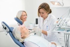 Female dentist examines an older patient's teeth with instruments while a companion waits in the bright clinic.