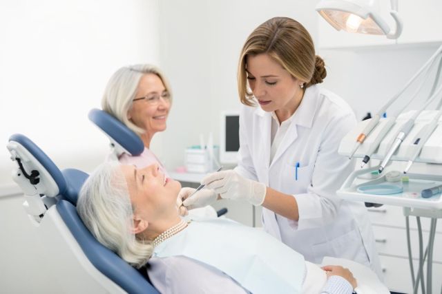 Female dentist examines an older patient's teeth with instruments while a companion waits in the bright clinic.