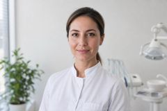 Female dentist in clinic wearing white coat, dental chair and light visible behind.