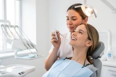 Dental patient smiling in chair while a dentist examines her teeth with a hand instrument in a bright clinic.