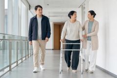 Elderly patient using walker in hospital corridor accompanied by two caregivers, suggesting orthopedic care.