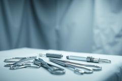 Surgical instruments laid out on a sterile surface. The image depicts stainless steel tools like scissors, forceps, and retractors against a blurred blue background suggesting an operating room or medical setting. The cool tones and shallow depth of field create a professional, clinical aesthetic.