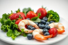 Healthy salad featuring fresh strawberries, blueberries, broccoli, lettuce, and cooked shrimp on a white plate. Vibrant red, green, and blue colors highlight a nutritious and delicious meal. Food photography, healthy eating, seafood salad.