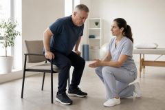 Clinician assisting older man rising from chair in a calm rehabilitation clinic setting.