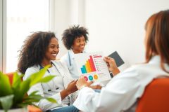 Two smiling women in white coats review a graph together in a bright, modern office setting. The image conveys teamwork and data analysis.